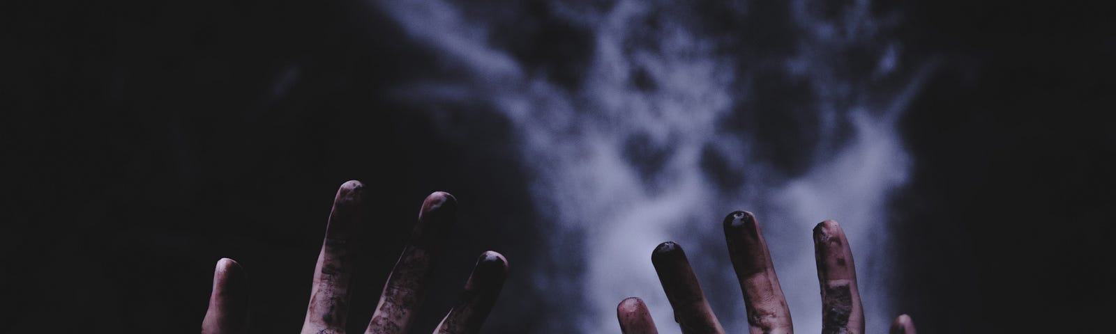 Photo of two hands in front of the camera, palms facing the camera with fingers splayed open, thumbs out and pinky fingers close together. The palms and fingers are dirty with a black-brown substance. Each wrist wears a string bracelet. The background is dark and blurry, with a tumultuous waterfall in the center of the image, well behind the hands.