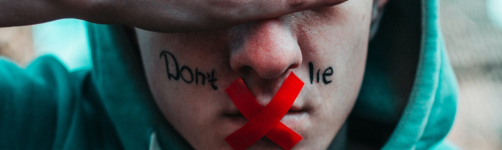 A male in a green hoodie covers his eyes with his hands. His mouth has a red X taped over it and the words “Don’t lie” are written on his face.