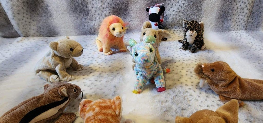 Photo of eight Beanie Babies facing a colorful ram Beanie Baby being humped by a rabbit Beanie Baby on a gray and white background. The ram looks stressed out.