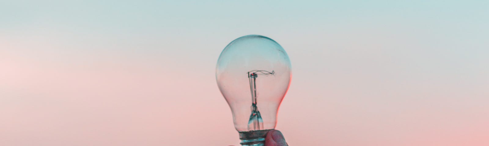 A hand holding up a light bulb in front of a blue and pink sky.