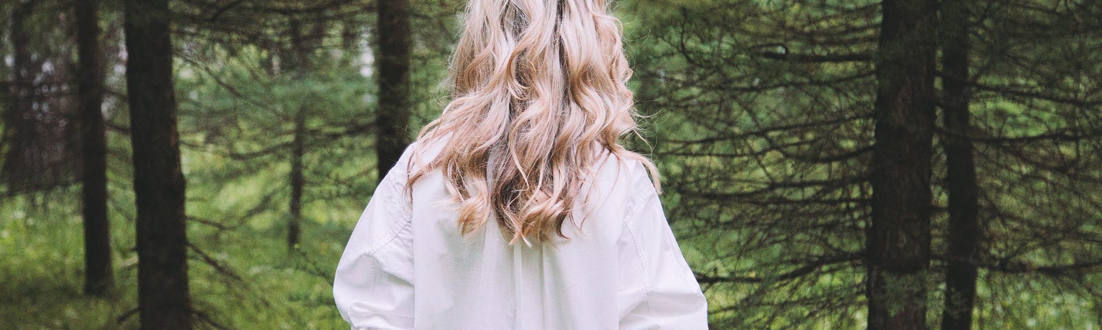 The back of a blond woman walking into the woods. She has a long while blouse and blonde hair.