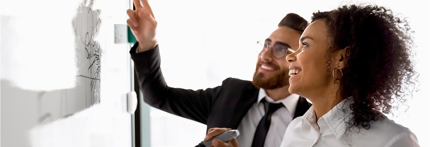 Male and female colleagues smile while learning together, standing in front of a whiteboard.