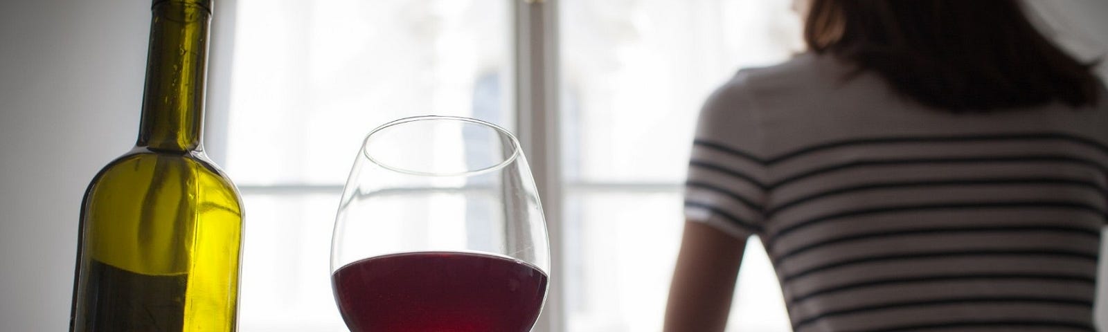 Image of a woman looking out a window in a darkened room with a wine bottle and glass with red wine in the foreground.