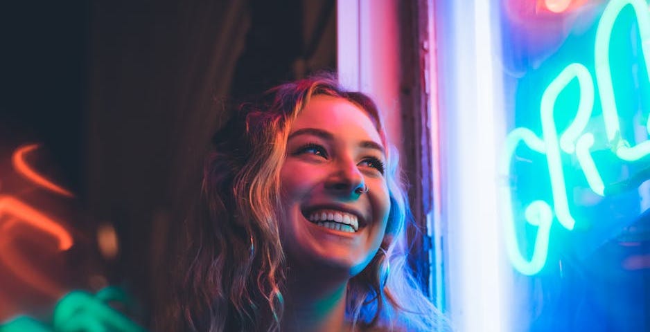Young smiling blonde woman looking up at a neon sign in the dark, her face bathed in rich, bright colours