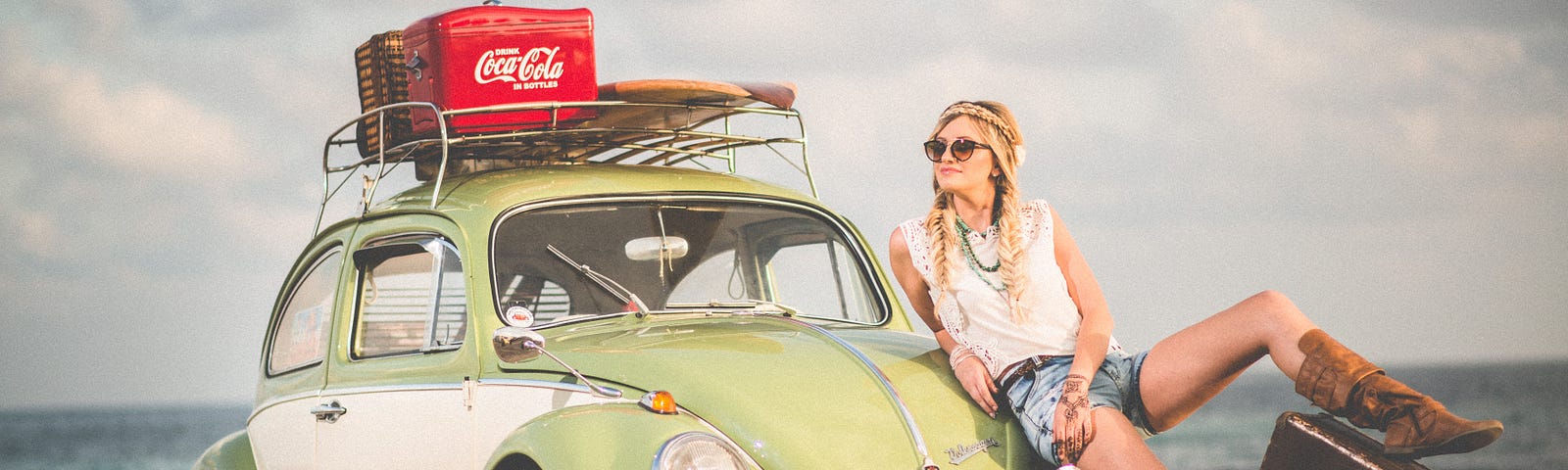 A blond woman with braids and sunglasses, one foot on a suitcase, leaning against an old VW bug, with a surfboard and boxes of Coke on the luggage rack. The ocean is in the background.