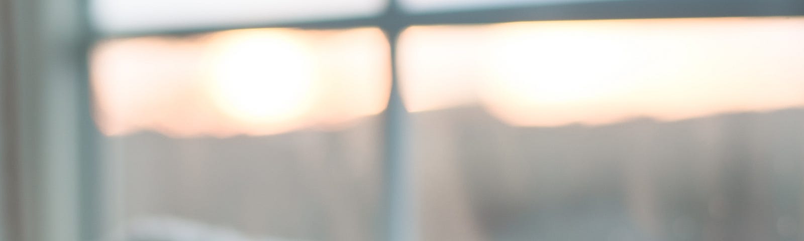 A small white coffee mug sitting on a pile of soft blankets. The backdrop is a blurry view through a frosted window, and it looks like a chilly winter sunrise is just beginning.