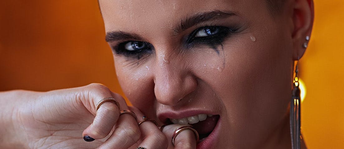 Young woman with dark eye makeup and dangling earrings biting finger and staring intensely at the camera