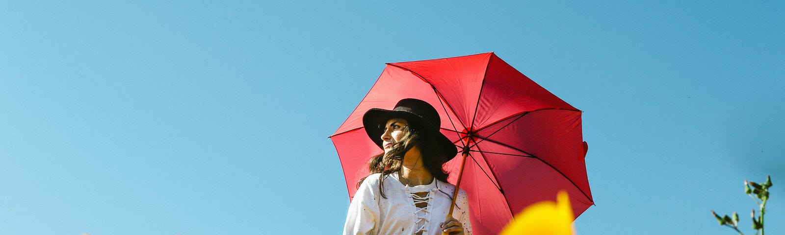 woman with a white dress and black hat, long hair flowing in the wind, holding a red umbrella and standing in a field of yellow flowers