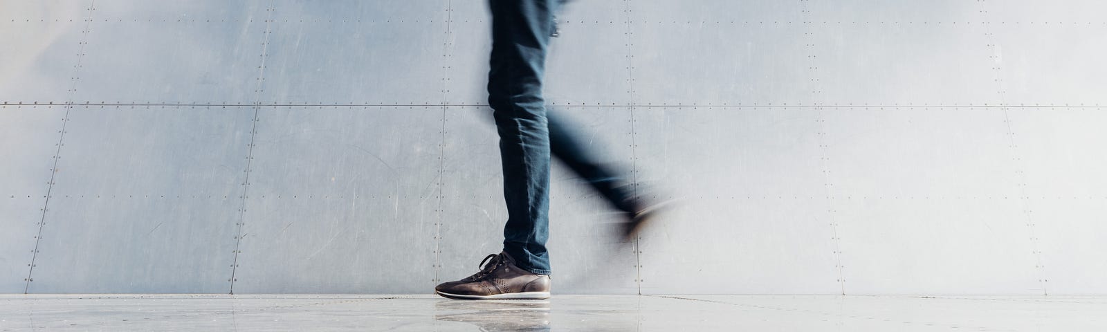 An individual walks from right to left. We see them from the abdomen level down, with the movement creating a slight blur. The person wears blue jeans, and there is a wall behind them.