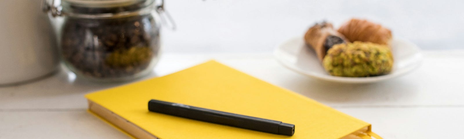 A yellow notebook with a black pen resting on the cover on a table with pastries and a jar of coffee on it.