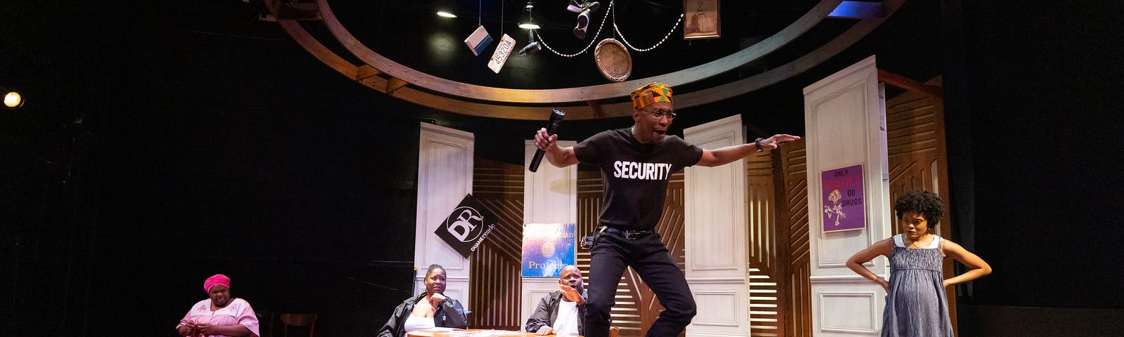 A man in a black t-shirt with the word SECURITY on it jumps off a chair while holding a flashlight and performing. His family members watch from the background.