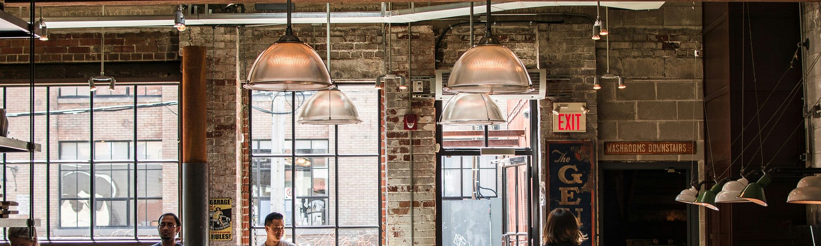 A busy, modern coffee shop with exposed brick walls, large metal-framed windows and vintage signs on the walls.
