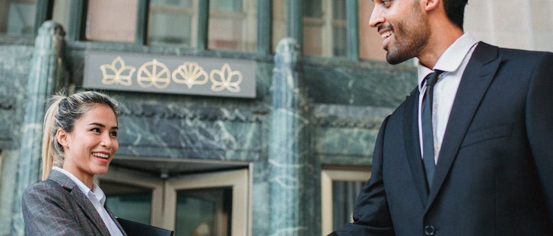 Male and female business people meeting outdoors in front of an old building and shaking hands. Both are professionally dressed and smiling