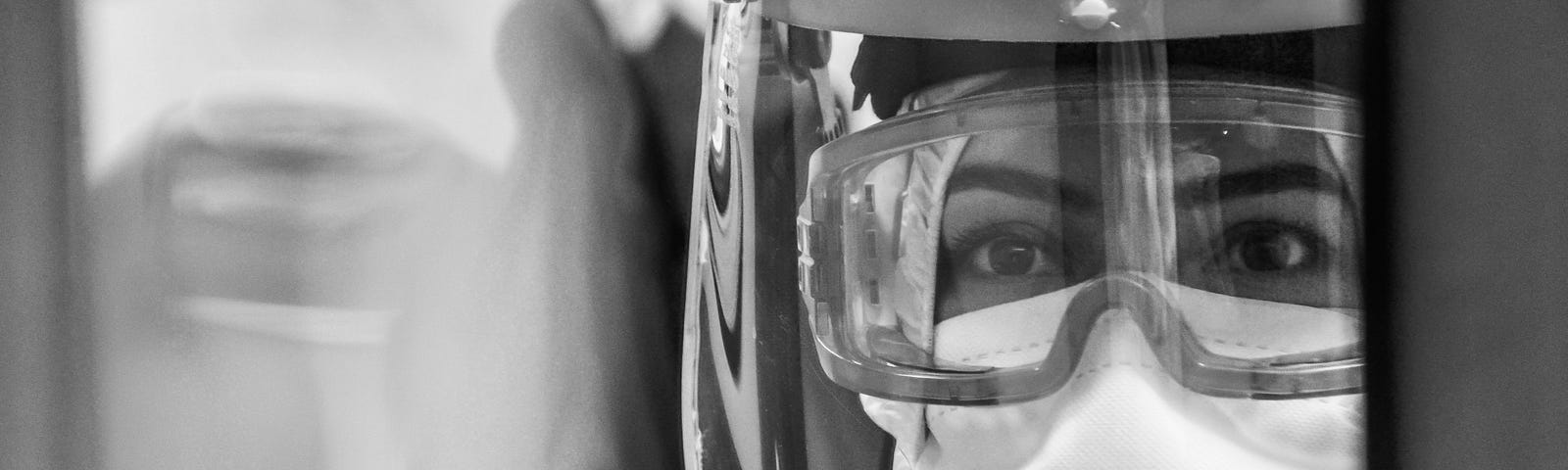 A healthcare worker in full protective gear, including a face shield, goggles, and a mask, looks through a window or screen with a focused and serious expression. The image is in black and white, highlighting the reflective surfaces and the clarity of the worker’s eyes.