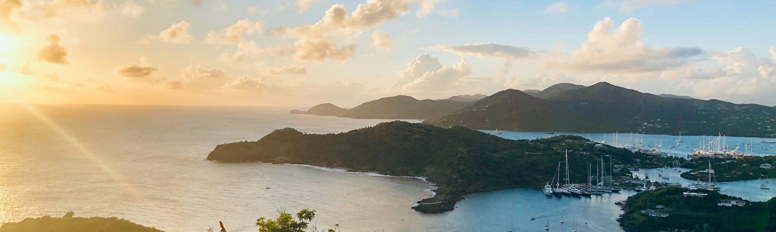 This image shows a beautiful coastal landscape during sunset. The sun is low in the sky, casting a warm glow over the scene. The foreground features rocky terrain with sparse vegetation, including some cacti. A bay with calm waters nestles below, where numerous boats and yachts are anchored. Rolling hills in the background complete this serene setting, and the sky is partially clouded, allowing rays of sunlight to peek through, enhancing the golden hour effect.