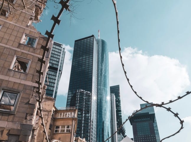 In the foreground is the debris of a destroyed building behind barbed wires. In the background, you can see posh skyscrapers and modern buildings.