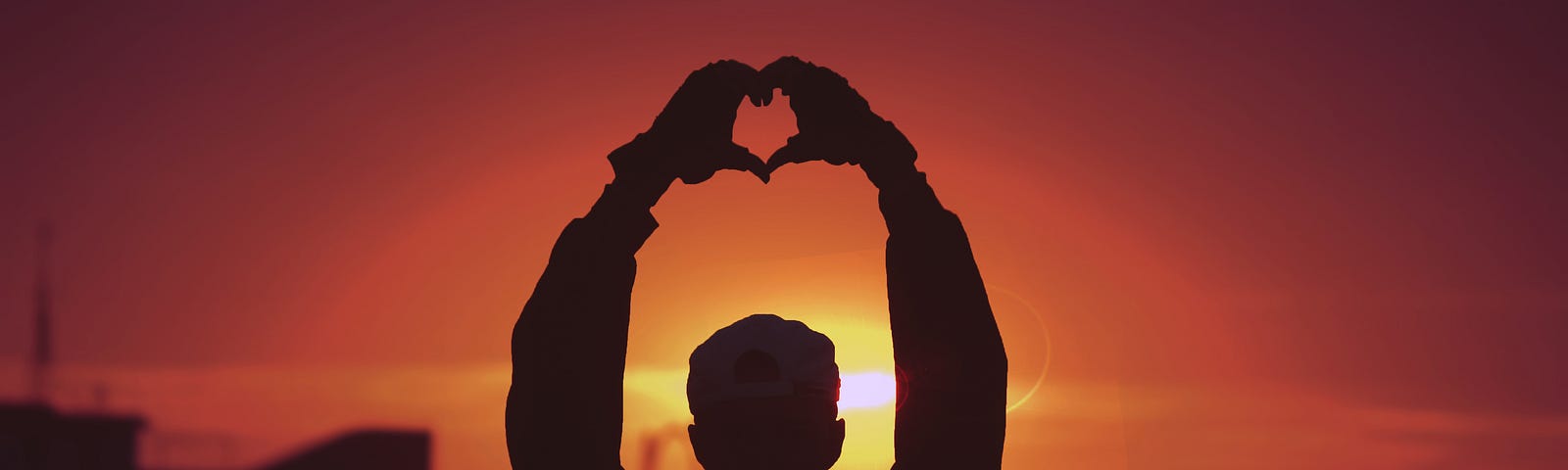 A person pictured at sunset. They are holding their hands above their head, using them to make a heart shape.