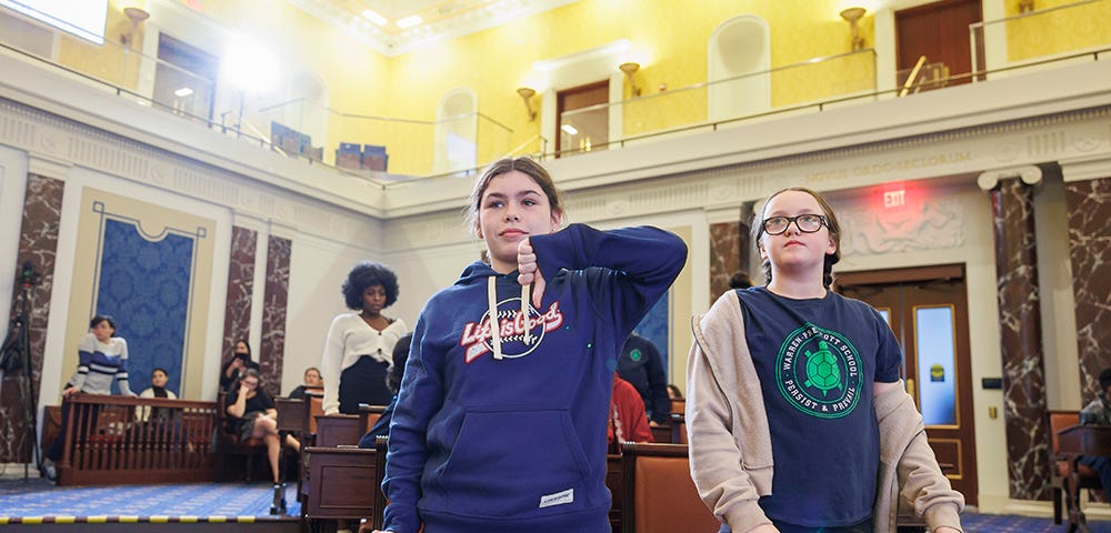 Photo of two middle school girls standing behind a desk in a replica of the U.S. Senate chambers. One girl shows her thumb pointing down for a “nay” vote.