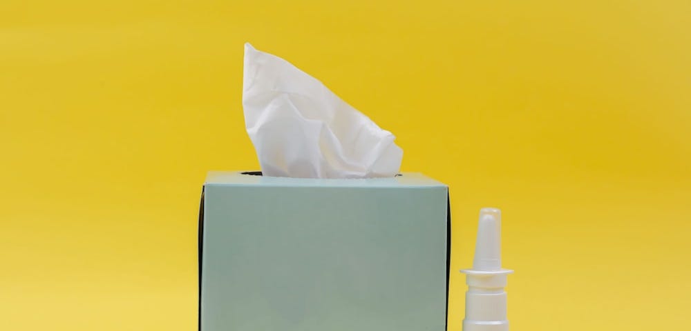 A green box of tissues and nasal spray in front of a yellow background