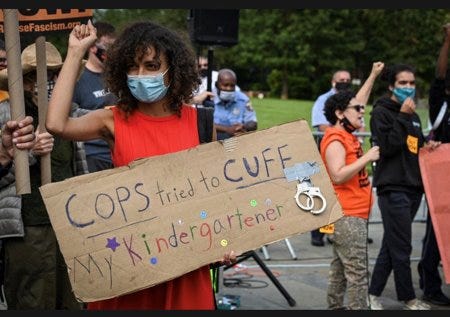 Photograph of activist Zoe Sturges, masked, at a protest. Her right arm is raised in a fist demonstrating solidarity, and she is holding a cardboard sign with handwritten text that reads: “Cops tried to cuff my kindergartner.” The word “kindergartener” is multi-colored, and a pair of cuff links are attached to the sign.