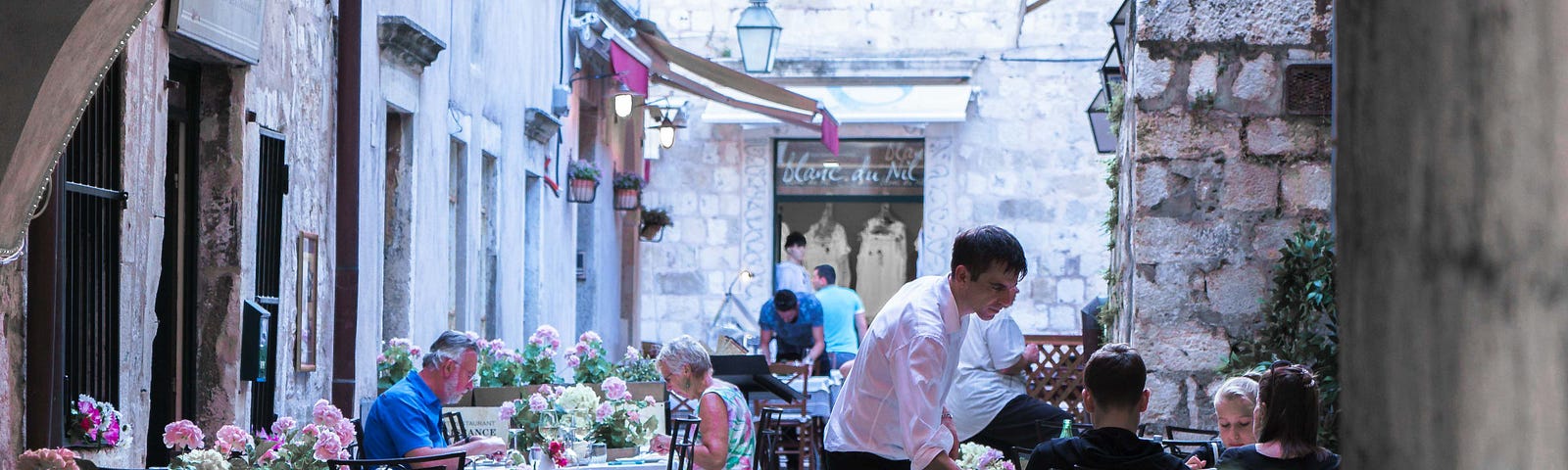 Tables with tablecloths and flowers lining coblestone street under a portico, waiter at one table with patrons.