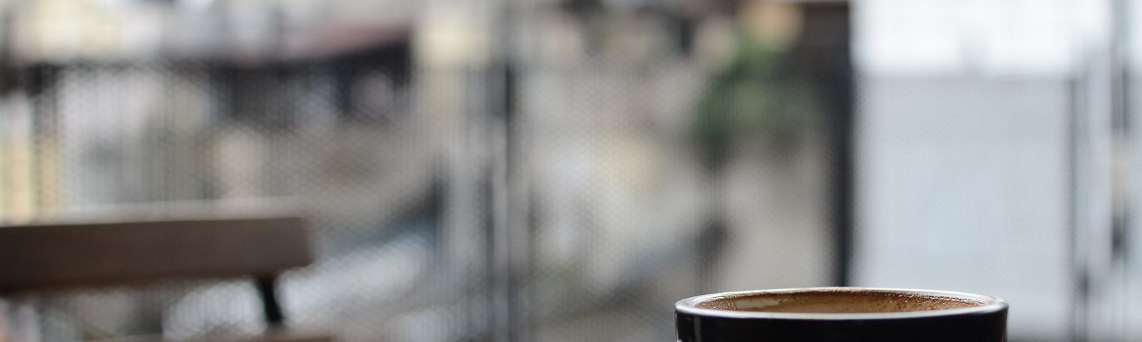 Photo of a single coffee cup sitting at the edge of a table in front of an empty seat, as if it’s for a ghost.