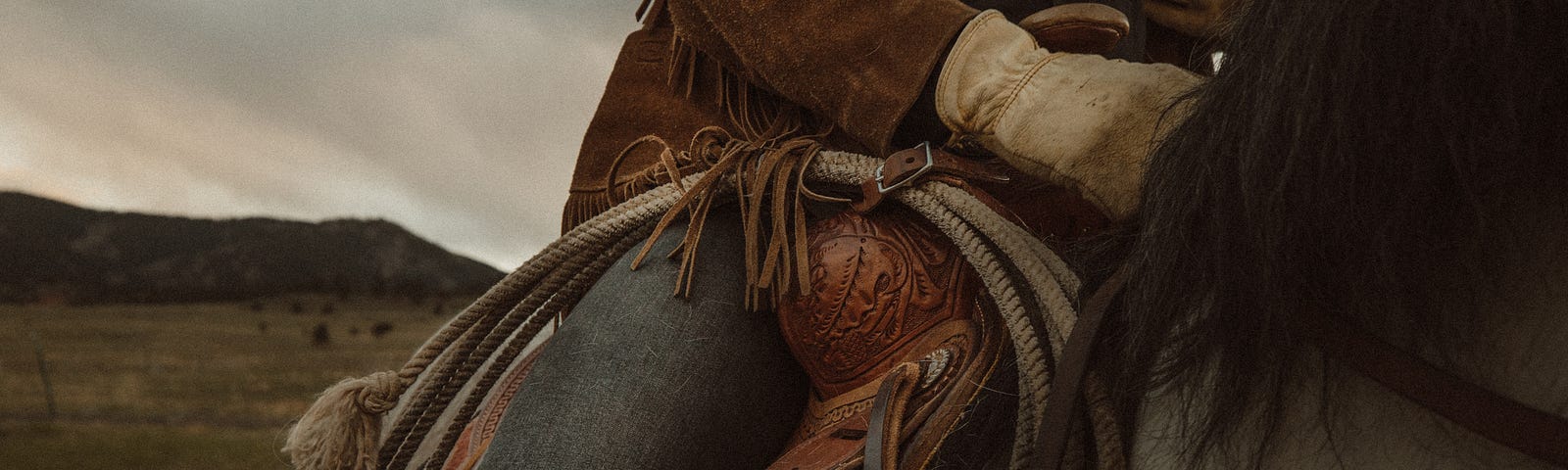 A close-up of a cowboy on horseback. The picture focuses on the beautifully-crafted saddle and a lasso attached to it. A bit of the grey horse and its shaggy black mane are visible. So are the cowboy’s jeans and brown leather jacket. You can see the prairie in the background and a flat hill.