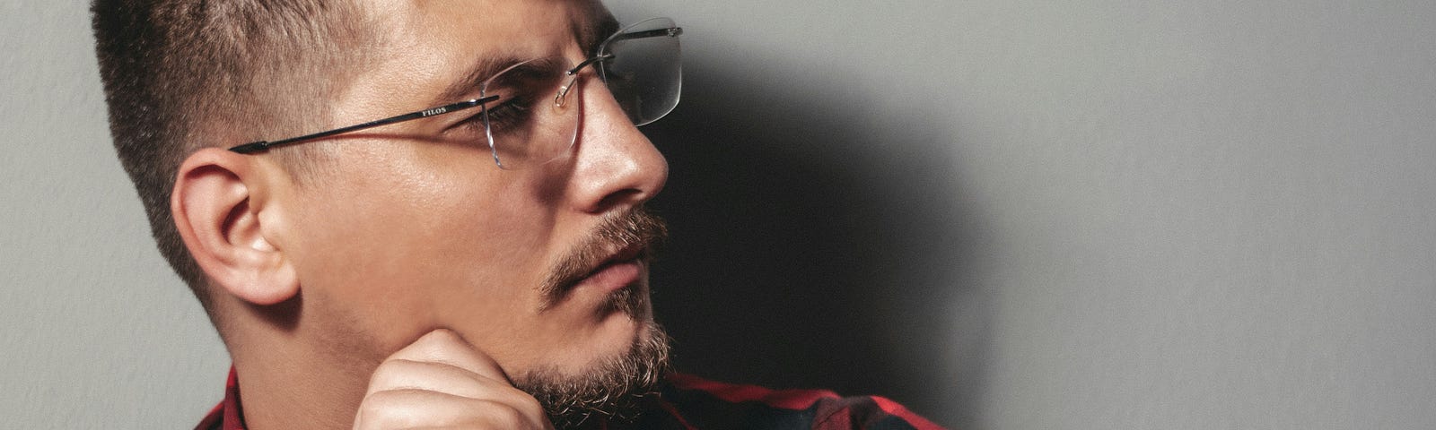 A young man with a mustache and goatee, a red plaid shirt and wearing glasses, looks left at something out of frame, in a “What the…” gaze.