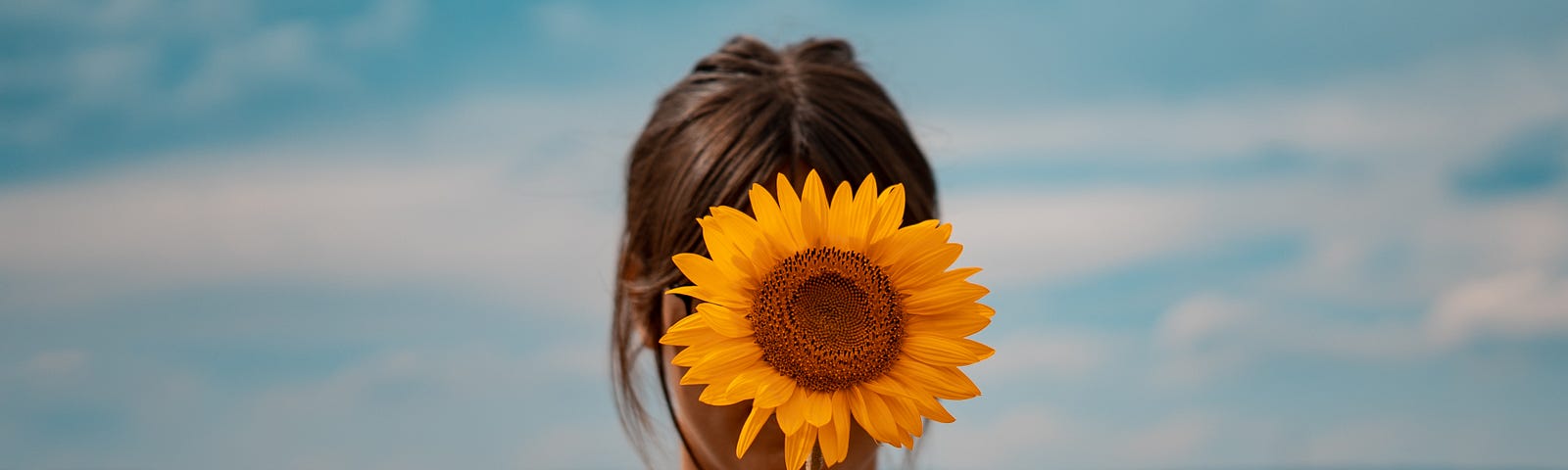 A young brunette woman faces us, her face obscured by a sunflower that she holds with her right hand. Colorectal incidence and mortality is rising among young adults.