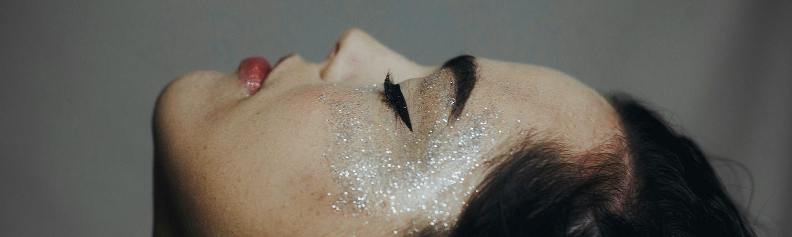 Profile view of a sensual woman with her head tilted back and eyes closed, with silver glitter artfully covering her cheek and temples.