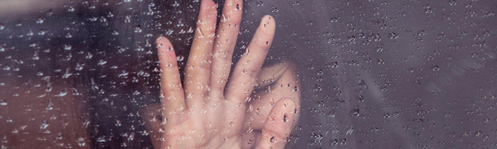 Distraught woman with hand covering part of her face pressed to a rainy window.