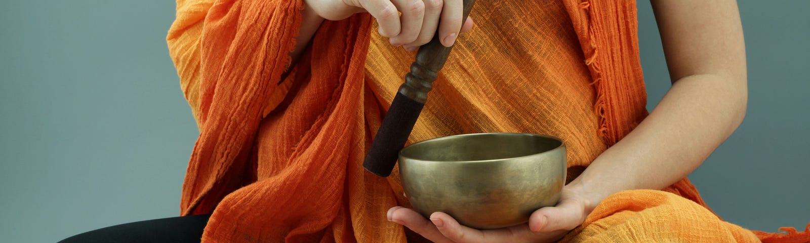 Torso only of someone sitting cross-legged, wearing black leggings and a wide orange shawl, holding a metal singing bowl in one hand and a wooden mallet in the other, apparently sounding them together.