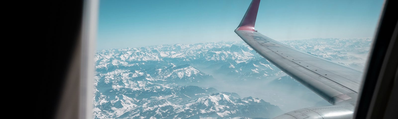 View of sky and snow-covered mountains under engine and wing of a jet window