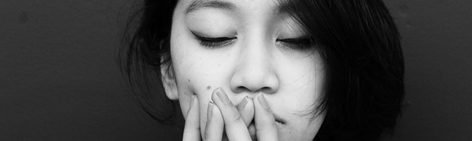 asian woman with her eyes clothes and hands over her mouth looking disappointed in black and white.