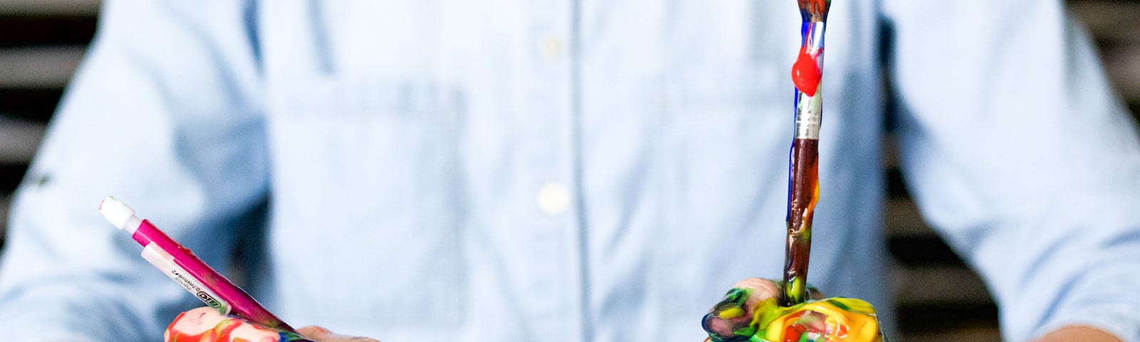 Man wearing a white shirt and holding a pencil and paintbrush in his hands and paint is on his hands