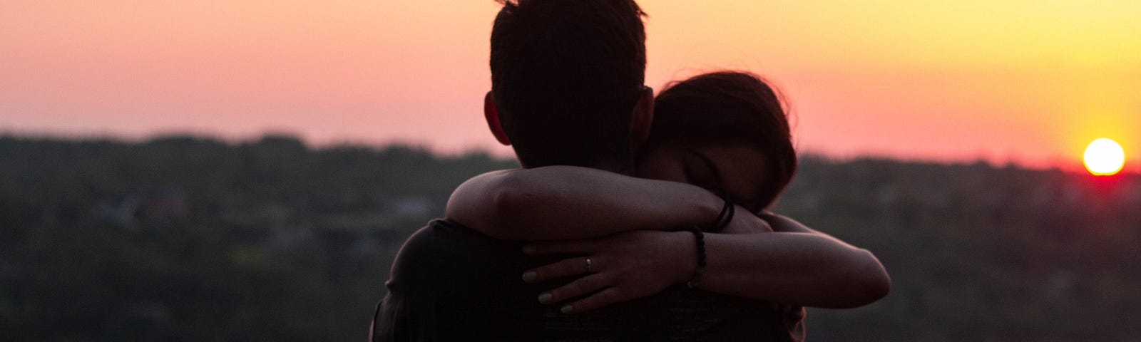 Two people hugging, silhouetted against the sunset. A lake behind them reflects the orange light.
