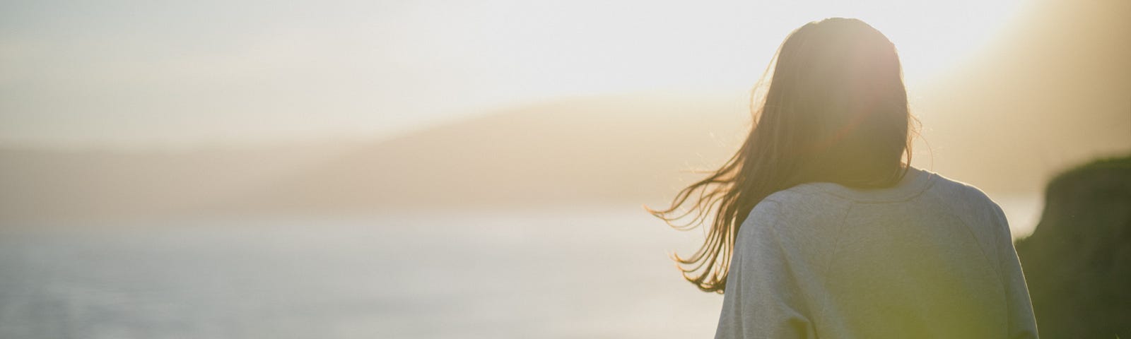 An attractive young lady, seen mostly from behind seems to be sitting on a clifftop looking at the see and the coastline as her hair blows gently in the wind.