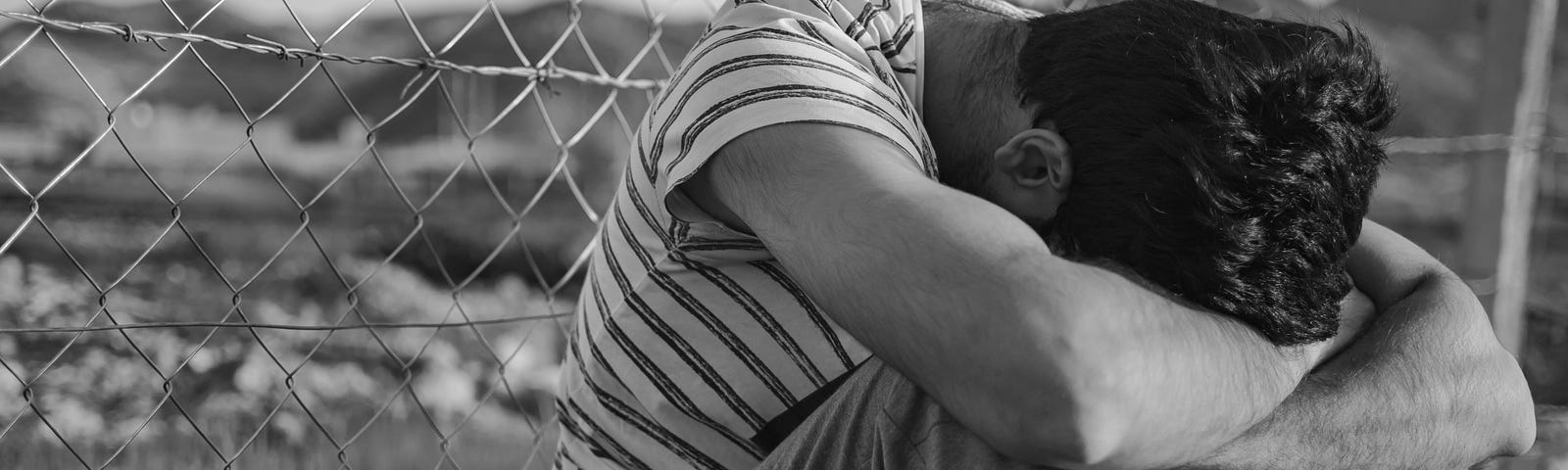 Man wearing striped t-shirt and jeans, sitting before chain link fenced pasture, arms crossed over knees, head bowed. Black and white photo.