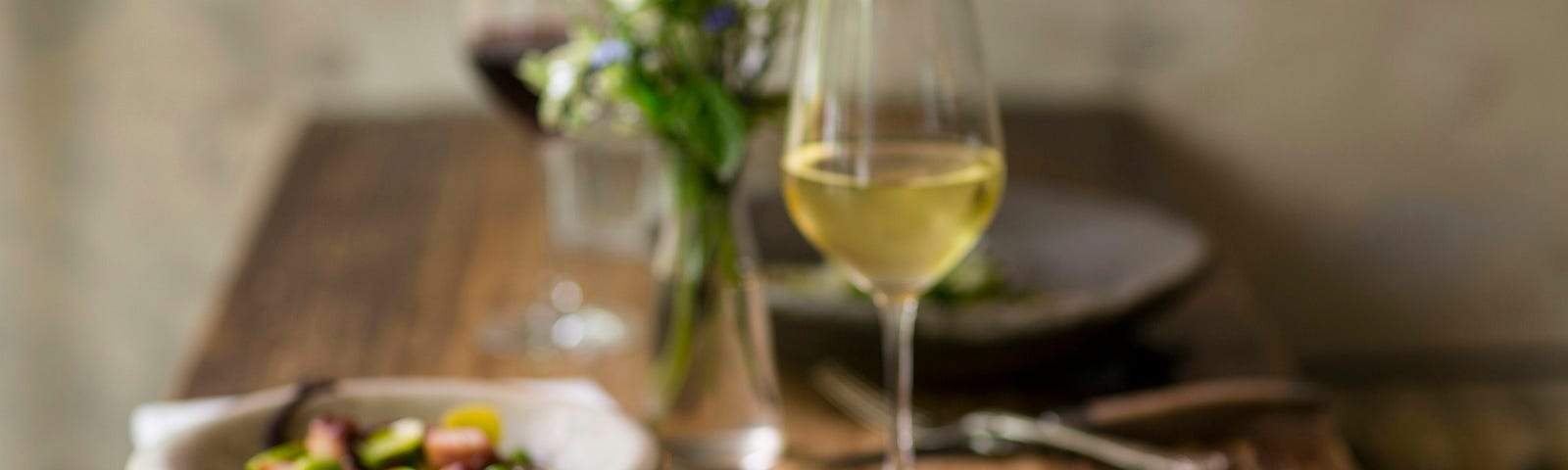 A glass of white wine and two elegant dishes sit on a wooden table.