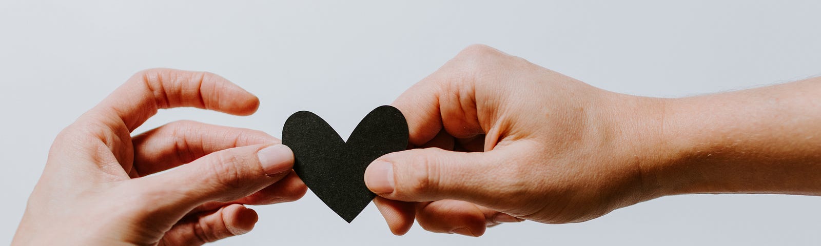 One hand reaches out from the left side of this image to recieve a black cutout heart being handed to them by somone else on the right. The background is a plain white.