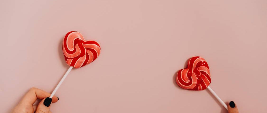 Two heart shaped lollipops held against a pink background