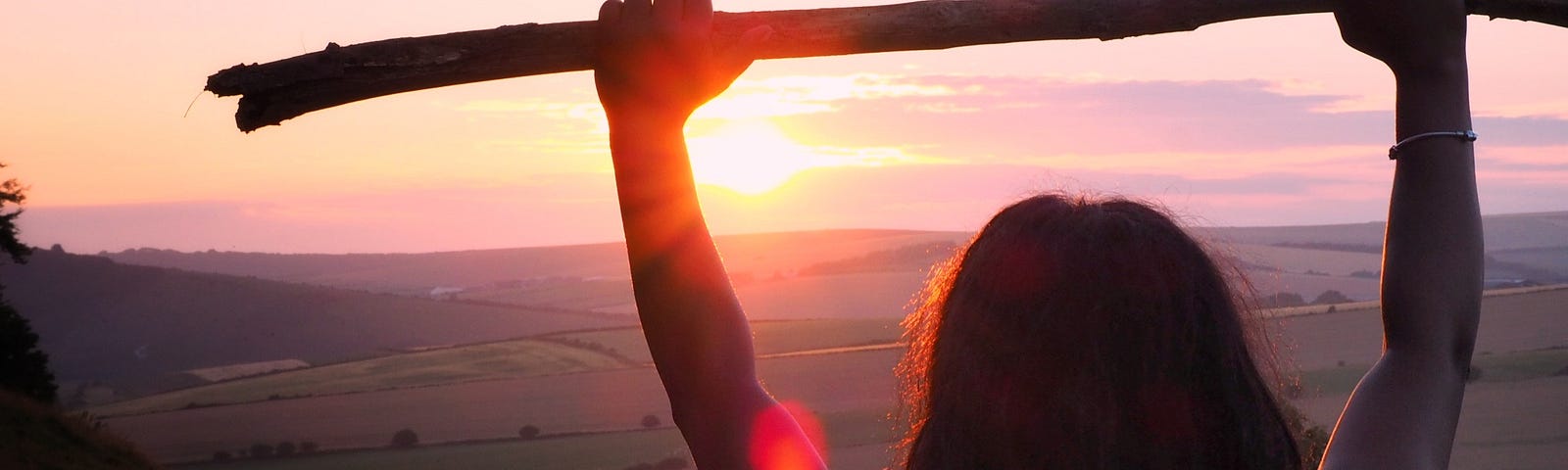 Woman raising a stick while facing a sunrise from a mountain top