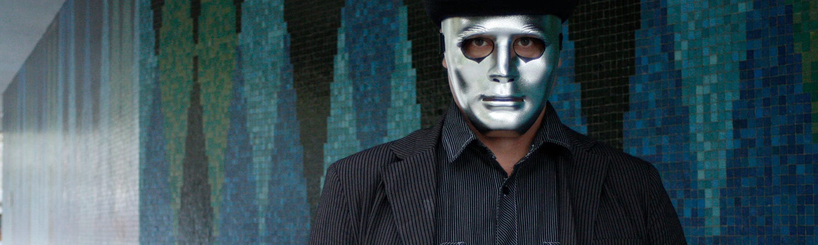 A man in black with a hat wears an ominous silver mask