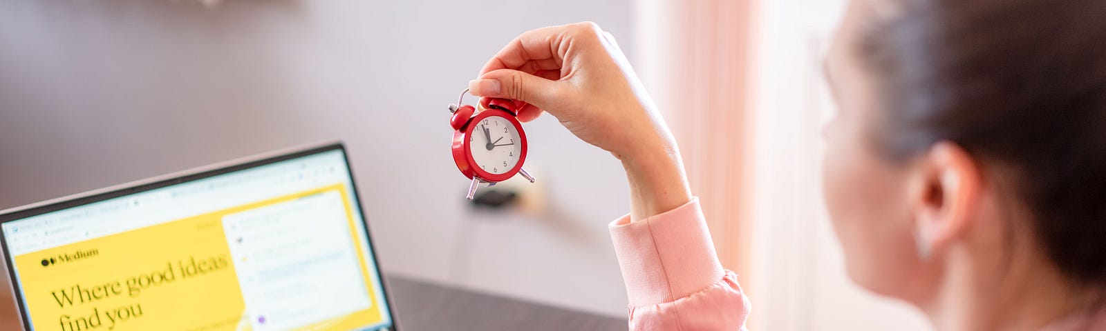 Woman holds up a small red clock (with a white face) as she looks at her portable computer. She sits in an office.