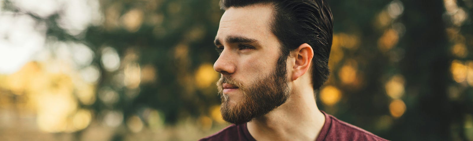 A guy in a purple-ish pullover, with dark hair and dark beard looking over his right shoulder as if looking for someone.