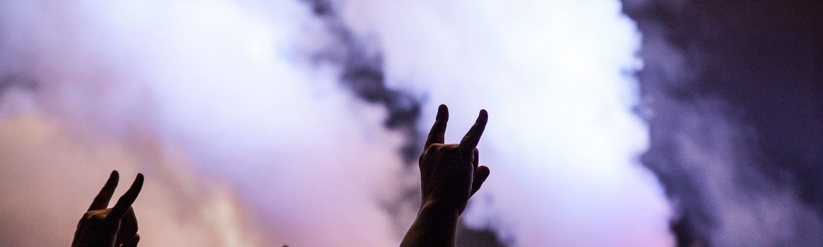 A group of people at a concert have their arms in the air, making devil horns against the haze of smoke from pyrotechnic displays.