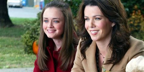 An image of the characters Rory Gilmore and her mother, Lorelai Gilmore, from the show “Gilmore Girls.” They are both white women with longish brown hair and blue eyes. Rory on the left is wearing a red coat and her hair pulled back from her forehead. Lorelai is wearing a camel coat and a necklace and has her hair swept back. They are smiling at something to the left off-camera.