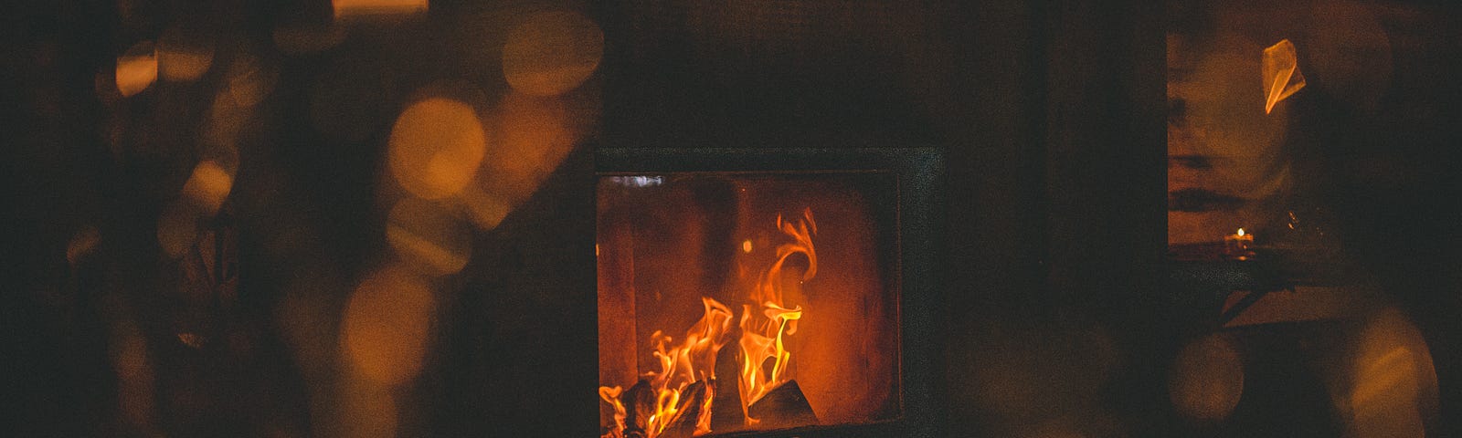 A fireplace with a crackling fire in a dark, timber/walled room. Only the fireplace in the centre of the picture is in focus. Around it are blurred golden lights that look like reflections of the fire in glas.