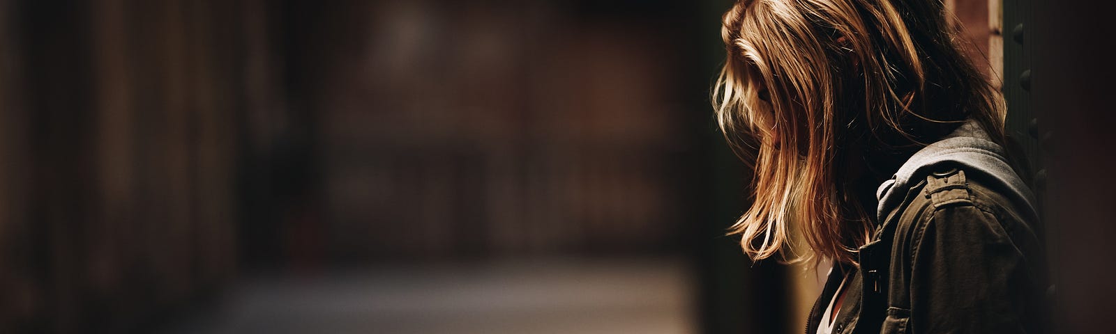 A young girl is leaning up against a wall in a blurred alleyway. The feeling is loneliness and shame. Her head is hanging down and her blonde hair is covering her face.