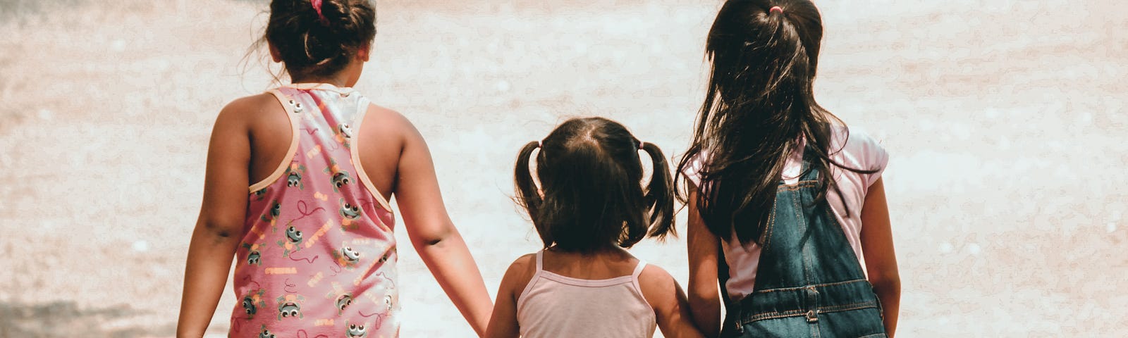 Three young children holding hands and facing the ocean, about to touch their toes in the water.